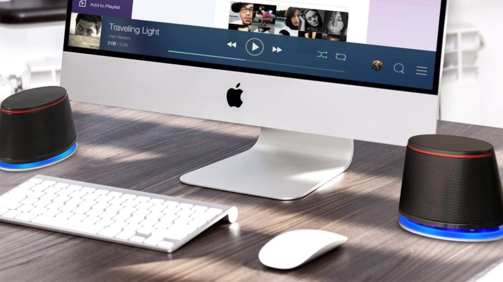 An Apple computer sits on a desk next to compact round computer speakers and a wireless keyboard.