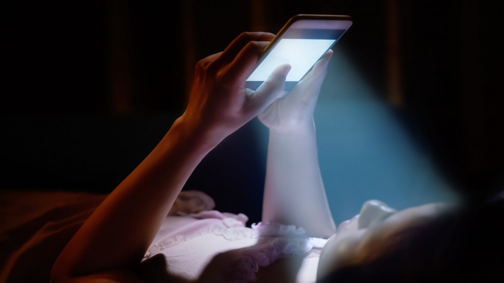 A young woman shining blue light on her face with a smartphone in bed